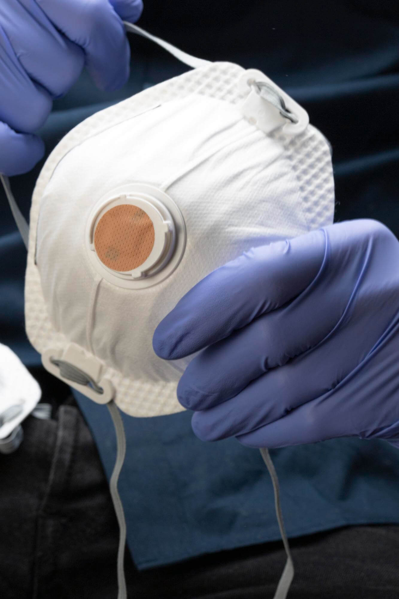 Lähikuva sinisissä lateksikäsineissä pidetystä valkoisesta hengityssuojaimesta, jossa on ruskea venttiili, tummansinisen taustan edessä.