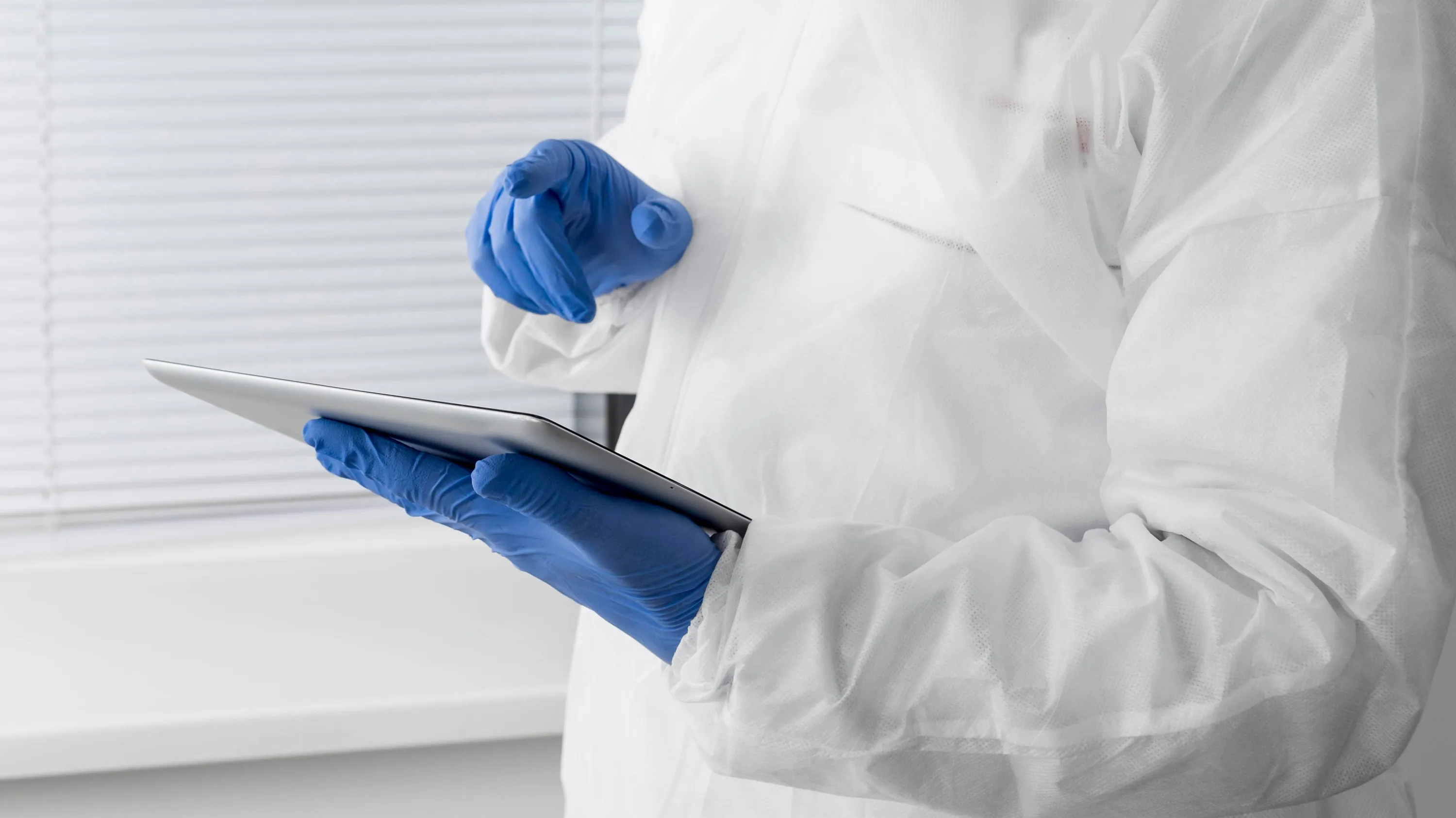 Kuvassa on henkilö suojapuvussa ja sinisissä käsineissä, joka pitää tablettia. Taustalla on valkoiset sälekaihtimet. Valo on kirkas ja tasainen.